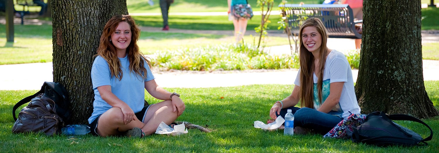 Two students seated in the shade of large trees in the central mall area of the campus